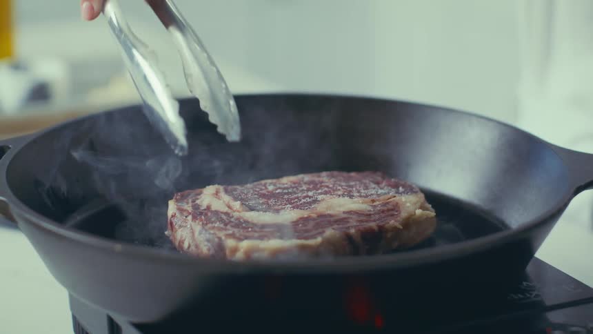 A thickcut steak sizzles and smokes in a castiron pan