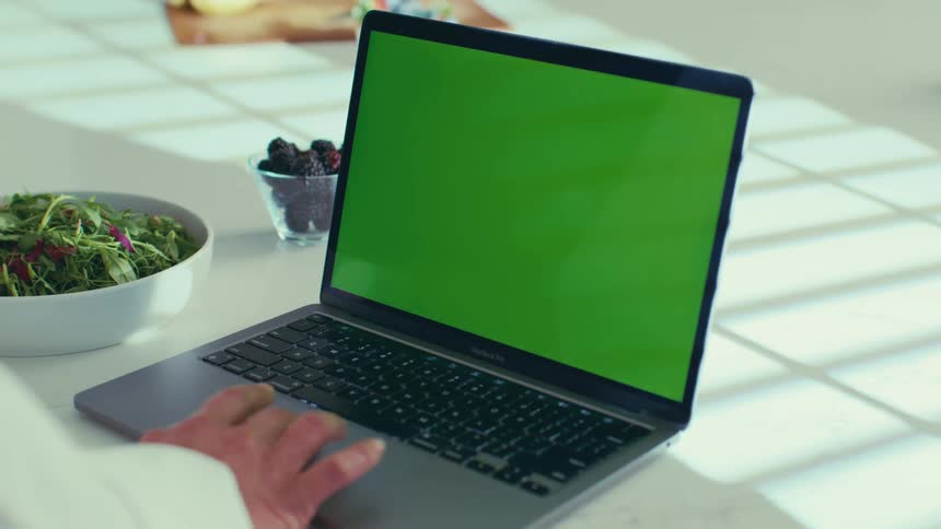 A woman typing on a laptop with a green screen while sitting at a kitchen counter with a bowl of salad and berries