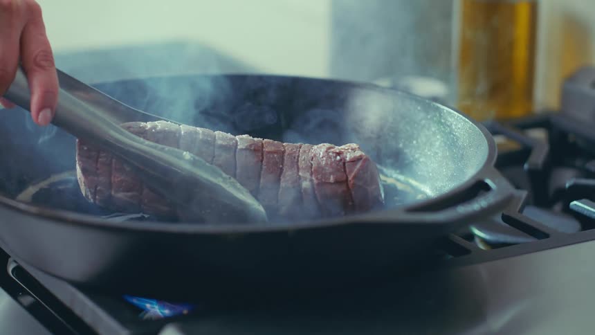 A large piece of meat being seared in a hot castiron pan on a stovetop with steam rising from the pan
