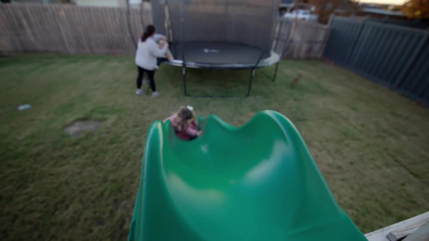 Little girl on a playground going down the slide and a woman in the background next to a trampoline