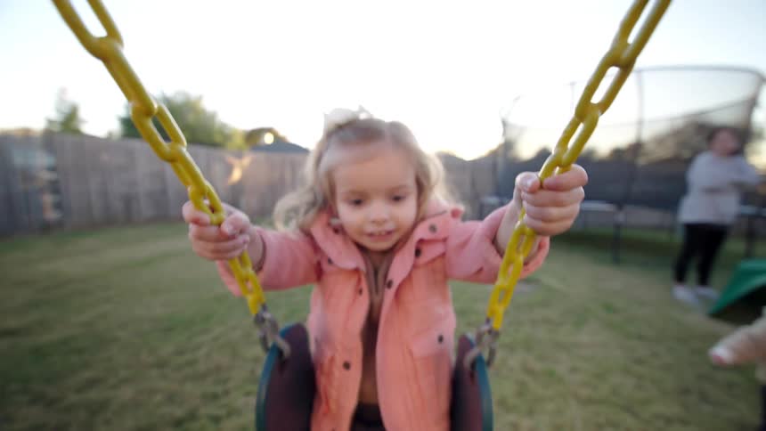 A happy girl in a pink jacket swings back and forth on a swing in a play area in the morning