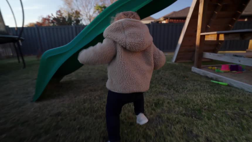 A little girl in a pink jacket runs around the backyard through a play area in the morning