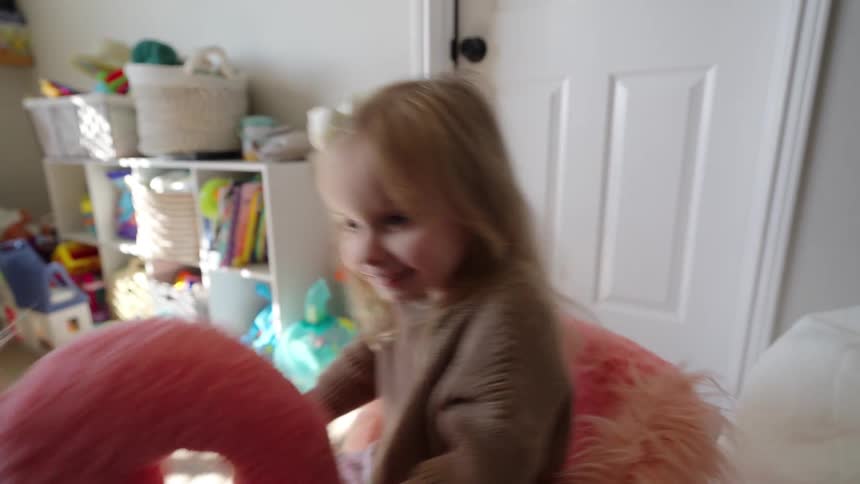A child is playing on a large plush pink flamingo toy in a room filled with various toys and storage baskets