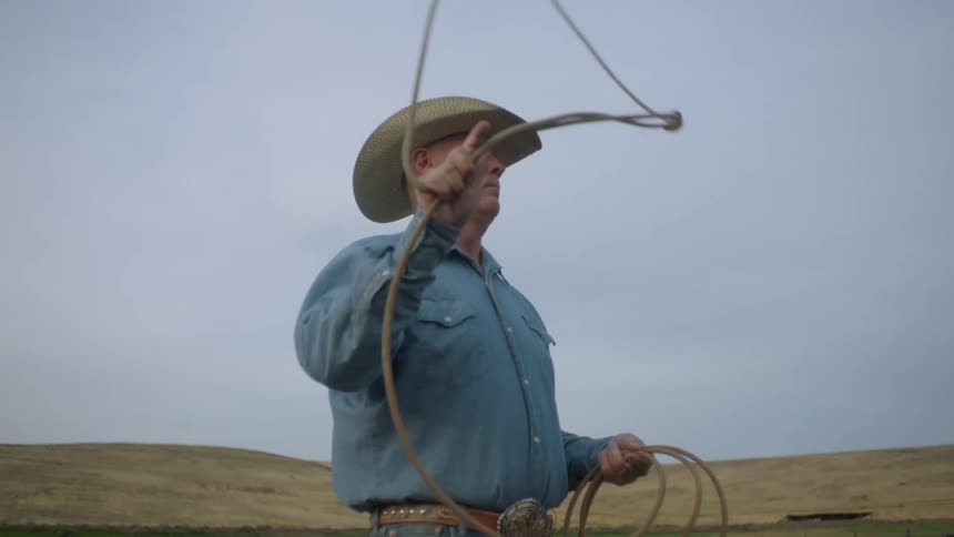An elderly man wearing a cowboy hat and blue shirt who is handling a lasso in an open field suggests a workrelated activity involving the lasso