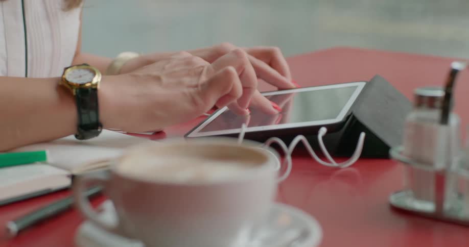 A womans hands using a tablet device while sitting at a table with a cup of coffee and a notebook