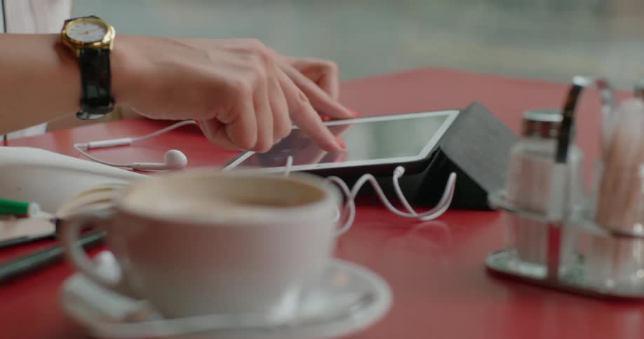 A woman wearing a wristwatch and white blouse sitting at a cafe table using a tablet while a cup of coffee and a notebook sit nearby