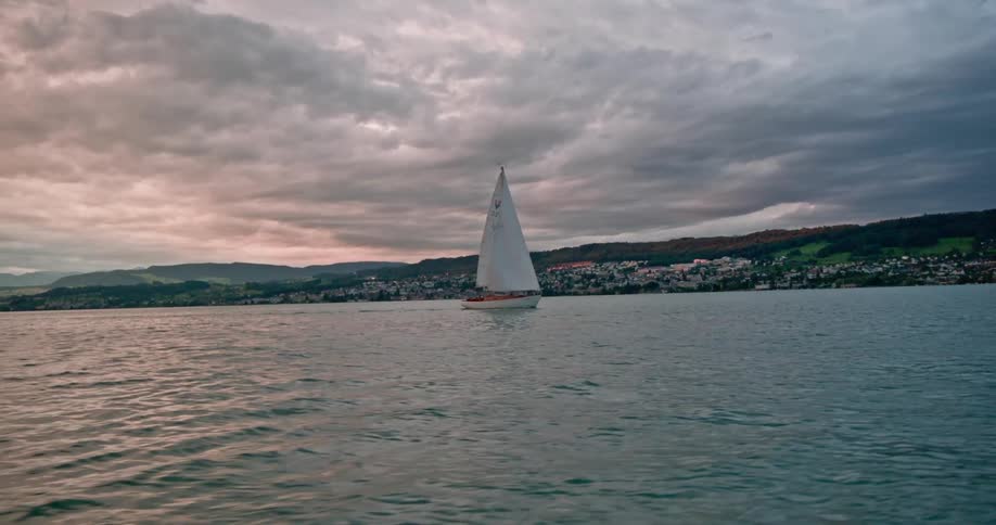 A sailboat gliding across a calm lake with a dramatic sky filled with dark clouds in the background and a picturesque coastal town visible on the horizon