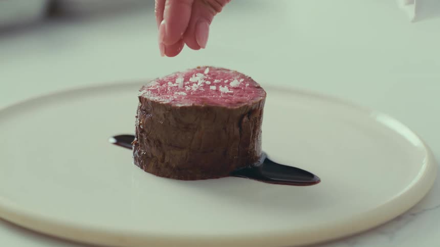 A chef seasons a perfectly cooked steak with a sprinkle of salt