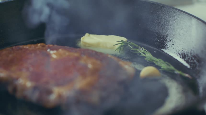 A steak sizzling in a hot pan with butter and rosemary creating a mouthwatering aroma