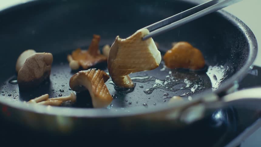 A pan with sliced mushrooms being cooked with a pair of tongs lifting one piece