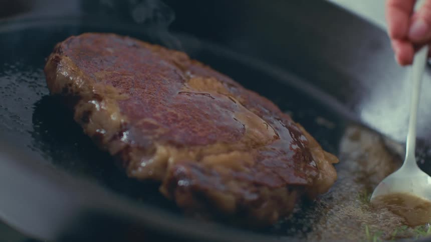 Hand with a spoon pouring a sauce on top of a steak being cooked in a frying pan