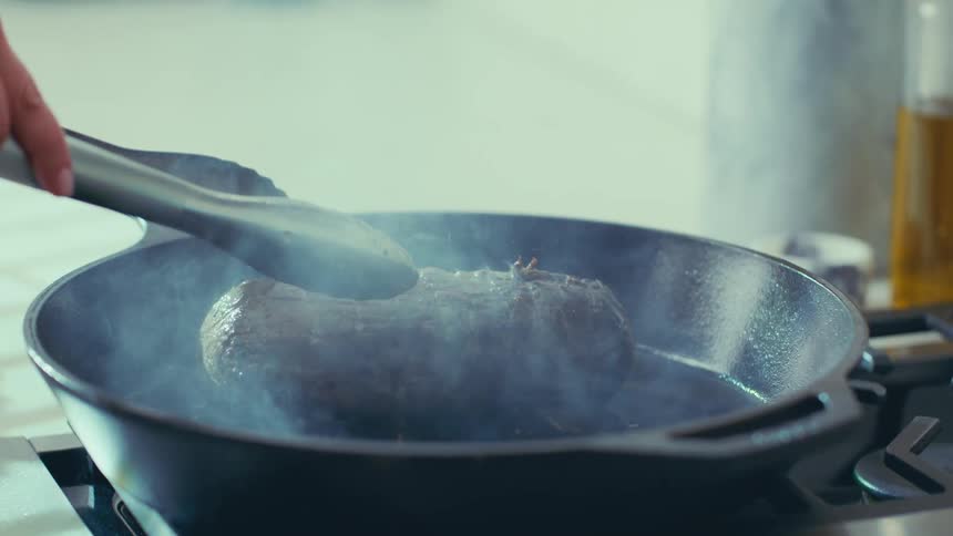 A pair of tongs flips a sizzling steak in a cast iron pan sending plumes of smoke into the air