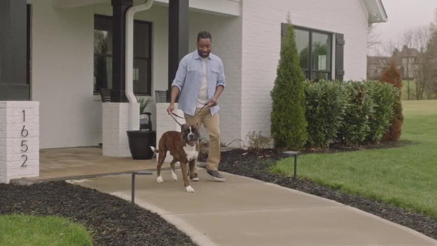 A man walking a dog on a leash on a concrete path near a white house