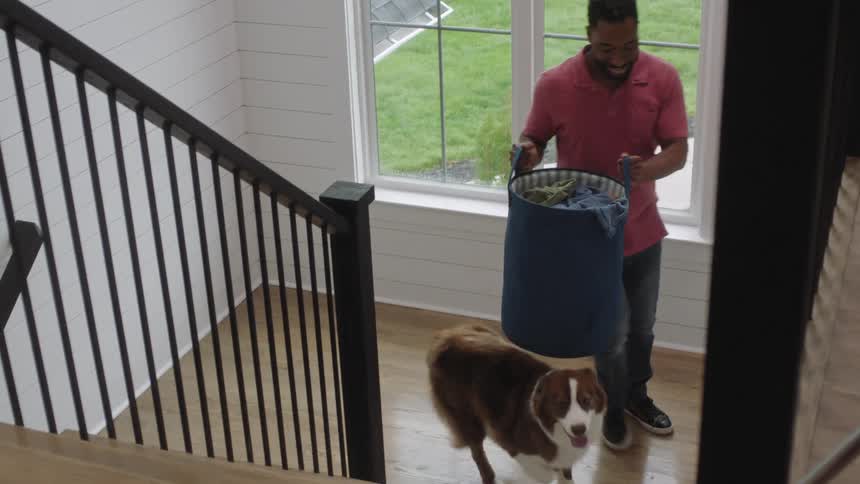 A man carrying a laundry basket upstairs is accompanied by a dog at home