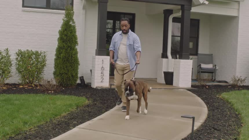 A man walks a dog on a leash as he comes out of a home and walks on the driveway