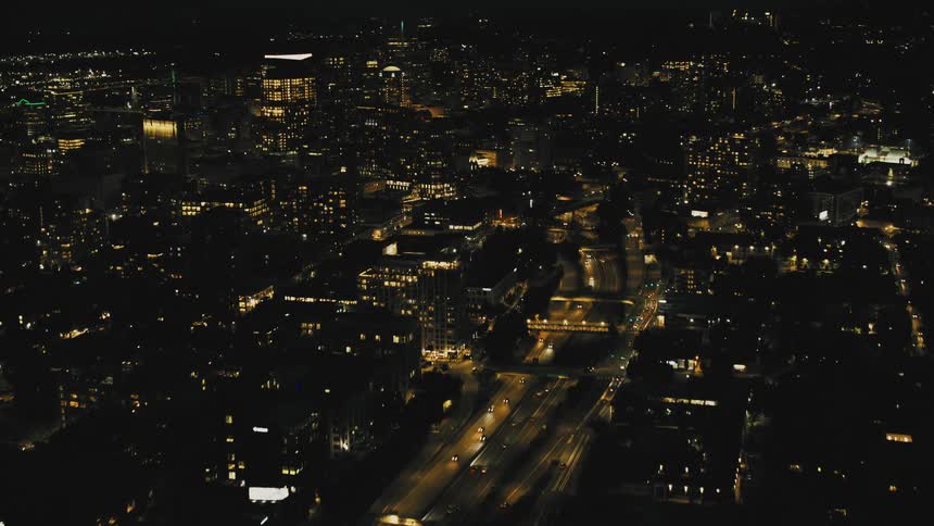 An aerial view of a city at night with a major highway illuminated by car headlights cutting through the densely populated area