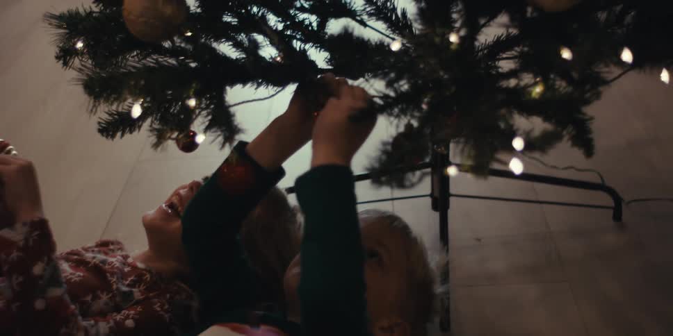Siblings lying on the floor slide on their backs under a christmas tree to place decorations underneath