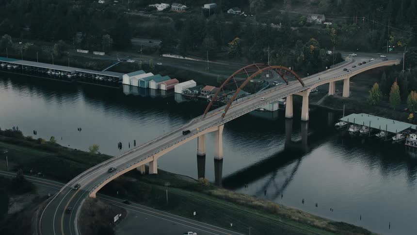 A large bridge with a red truss spans a wide river connecting two sides of a town where traffic flows along the bridge and boats are moored below