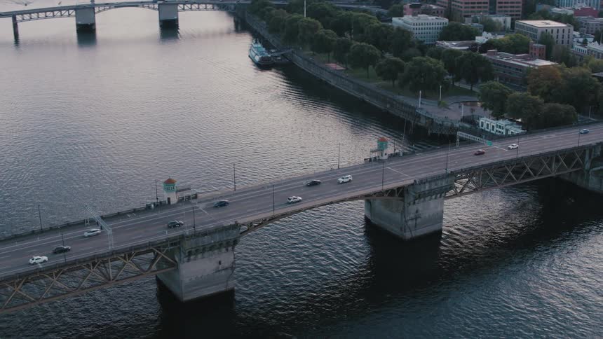 A suspension bridge that passes over a canal and there are several cars passing over the bridge on an asphalt floor