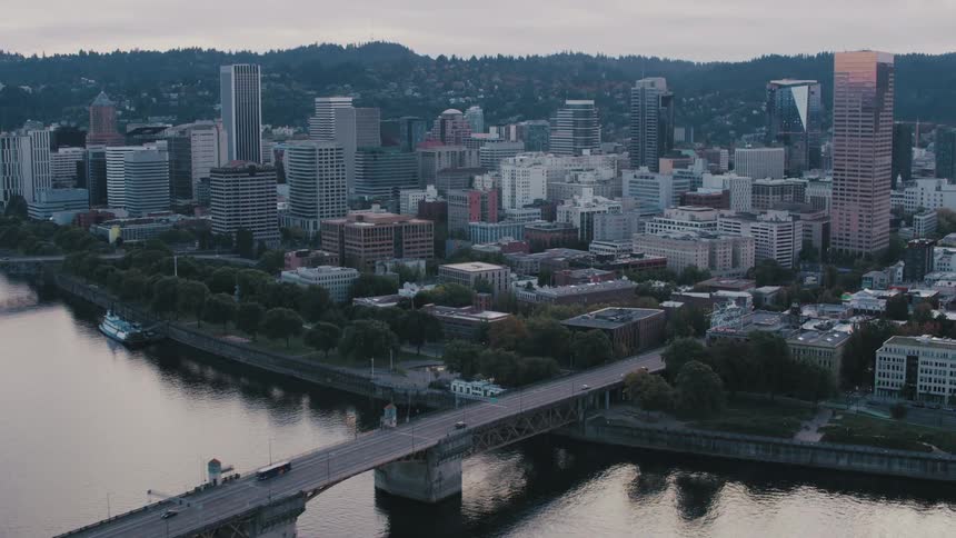 An aerial view captures a bustling city at dusk where skyscrapers pierce the twilight sky casting long shadows across the cityscape while a wide river winds through spanned by several bridges carrying traffic across its waters