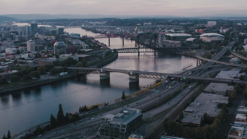 An aerial shot shows the city of portland oregon at dusk with the willamette river curving through the city multiple bridges crossing the river and a high rise building prominent in the foreground
