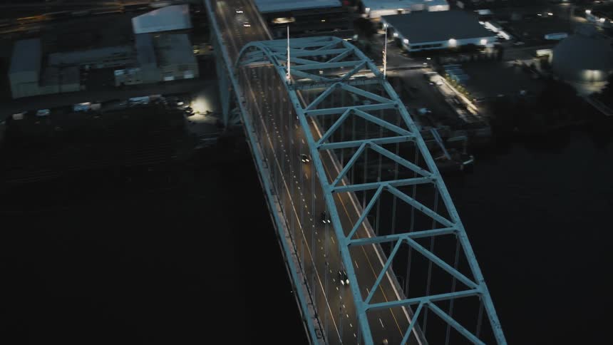 An illuminated bridge arches over a dark river at night with traffic moving along the road below
