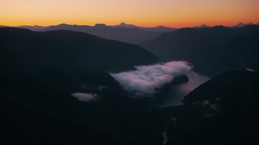 A mountain range is silhouetted against a vibrant orange sunset a river winds through the valley below partially obscured by fog