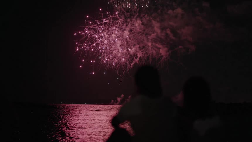 People sitting along the coast watching fireworks explode in the night sky over the water