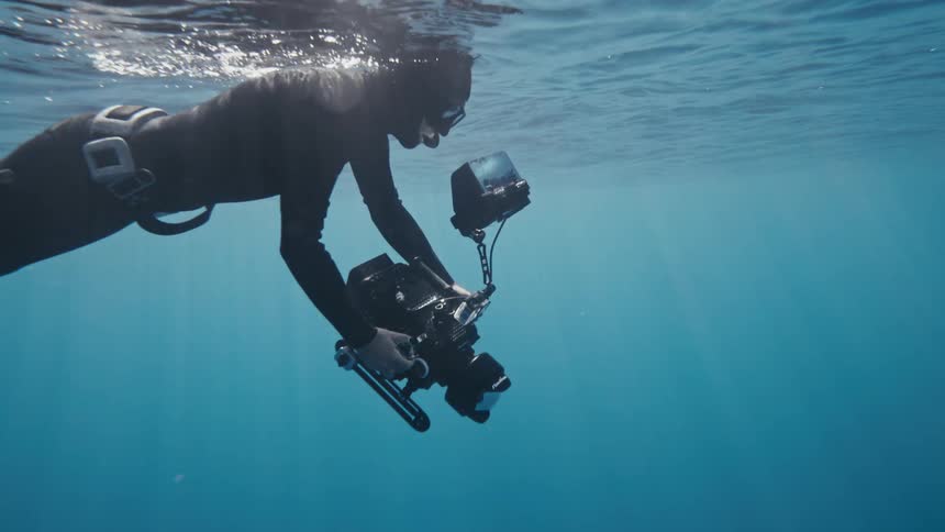 An underwater photographer wearing a wetsuit and diving gear is submerged in clear blue water holding a professional camera rig with a monitor and lighting equipment