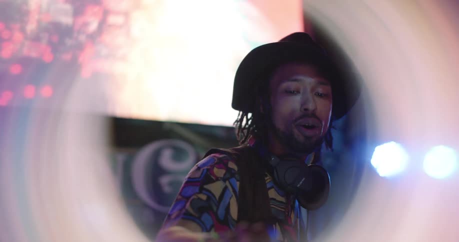 A man wearing a hat and headphones is shown in a close up as he performs or speaks in front of a colorful blurred background