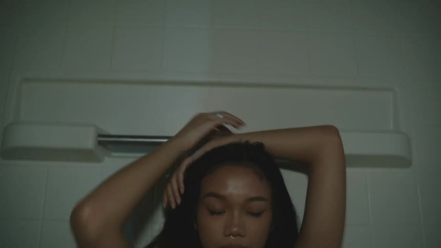A young woman stands under a dimly lit shower with her eyes closed one hand resting on her head and the other near her ear her serene expression framed by long dark hair against a minimalist backdrop of white tiles and a shower rack