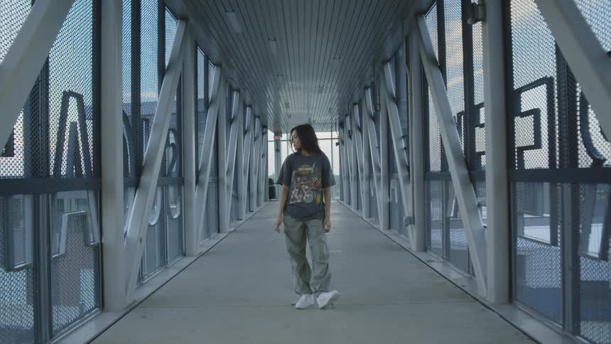 A teen is standing on a modern enclosed pedestrian bridge with metal and glass structure wearing casual clothing and white sneakers
