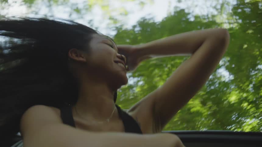 A young woman hangs out of the sunroof of a moving vehicle as her long hair blows in the wind