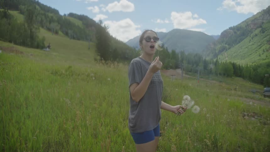 A woman blows dandelions in the valley with hills in the background