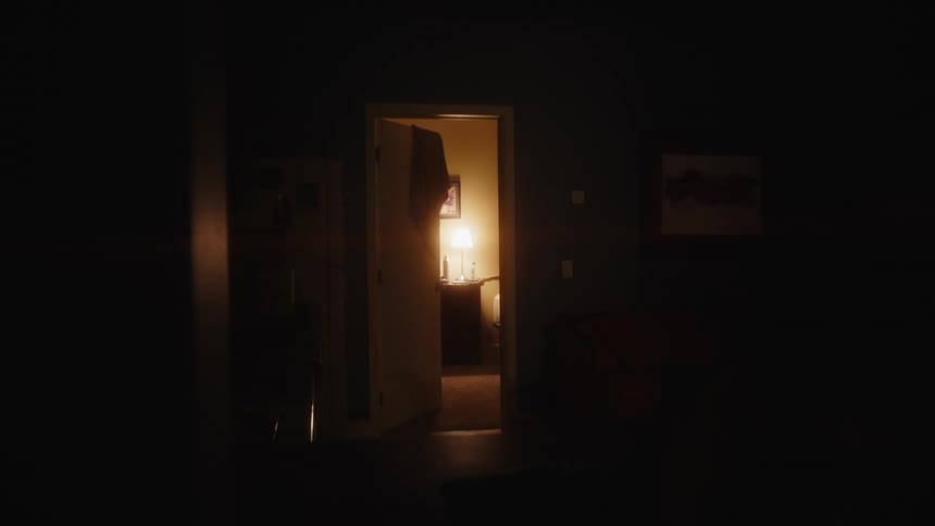 A dimly lit hallway leads to an open doorway revealing a brightly lit bedroom with a bed a nightstand and a framed picture on the wall