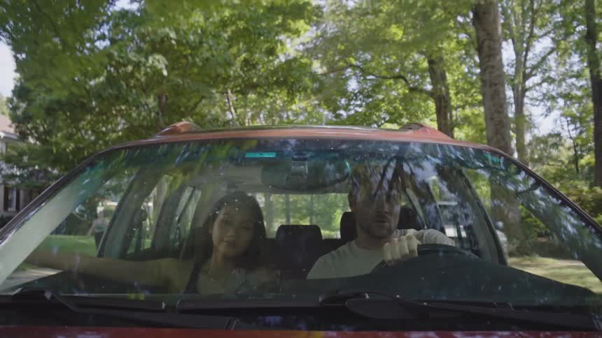 A husband and wife drive through the suburbs among many trees and scattered houses