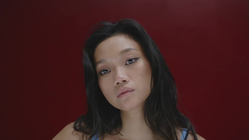 A woman with a nose piercing and black wavy hair looks around in front of a crimson red background