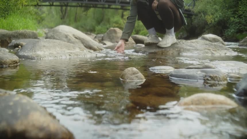 A person sits on a rock beside a shallow clear stream gently touching the water with their hand
