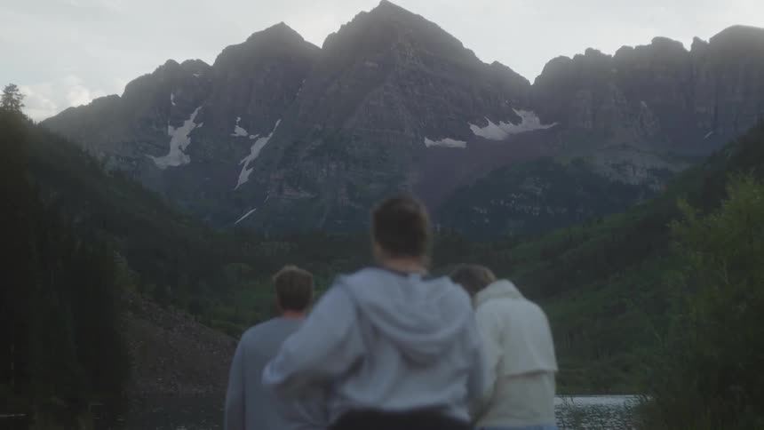 Three people stand near a lake looking at a large mountain range in the distance