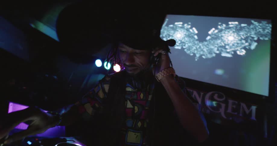 A dj wearing a hat and patterned shirt works at his mixing board in a dark club with flashing colored lights