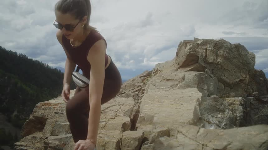 A woman wearing sunglasses and athletic attire is getting off a rocky ledge atop a mountain surrounded by rugged cliffs with a cloudy sky and distant forested hills in the background