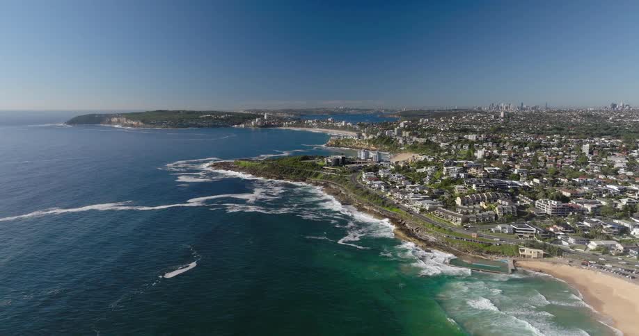 An aerial view of a coastal city with beaches stretching along the coastline a harbor filled with boats and a cityscape rising in the distance