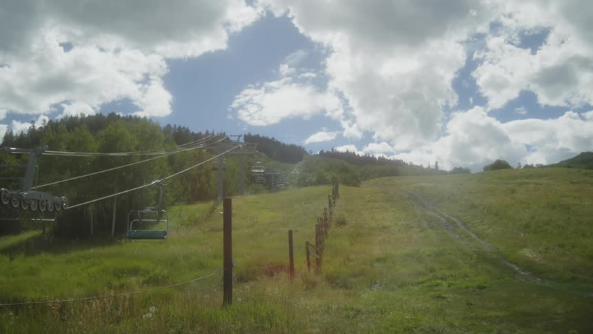 A bright sunny day at a ski resort with a chairlift ascending a green grassy slope white puffy clouds are scattered across a blue sky