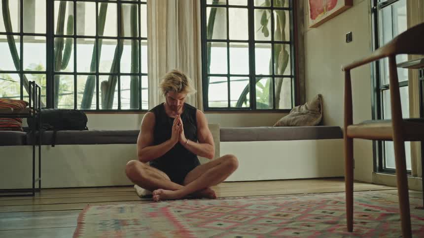 A man sits cross legged on a rug in a serene sunlit room with large windows palms pressed together in a meditative pose surrounded by minimalist furniture and plants