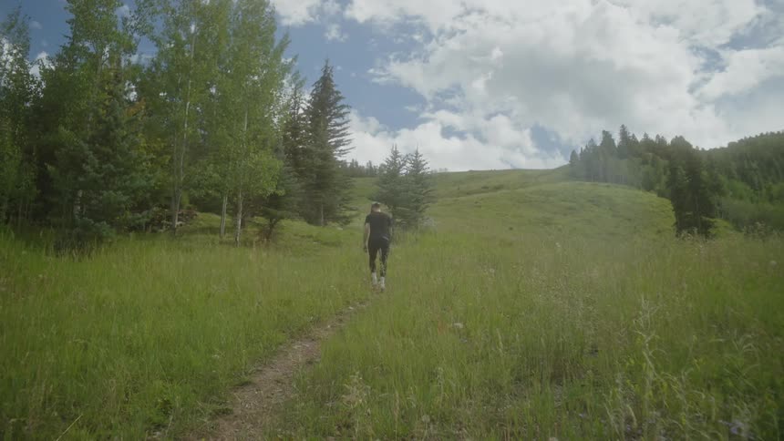 A man in black clothes is walking on a dirt trail in an area with lots of green grass in the environment