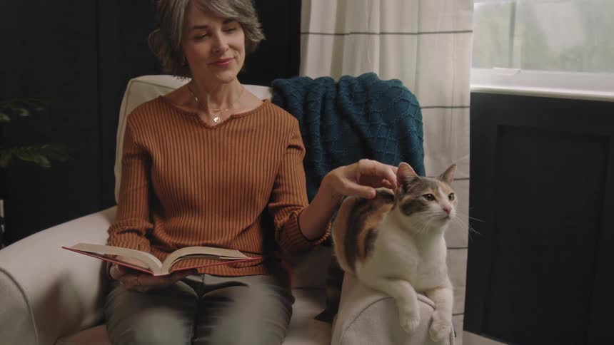Woman sitting in a chair reading a book as she pets her cat sitting next to her on the arm of the chair
