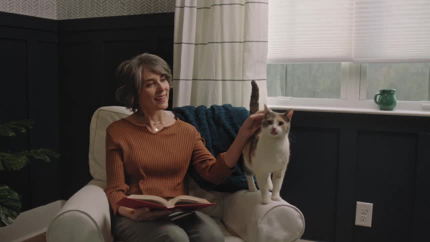 Woman sitting in a chair with a book smiles as her cat moves next to her on the arm of the chair and pets it