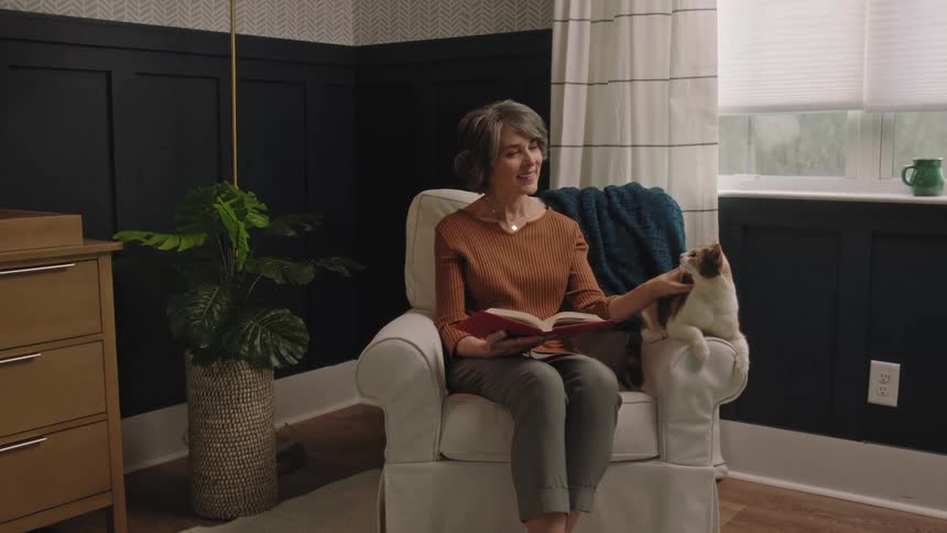Woman sitting in a chair reading a book as she pets her cat sitting next to her on the arm of the chair