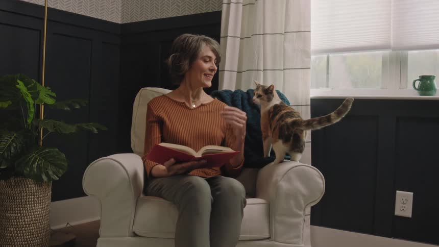 Woman sitting in a chair reading a book when her cat jumps in the chair as she smiles and pets it