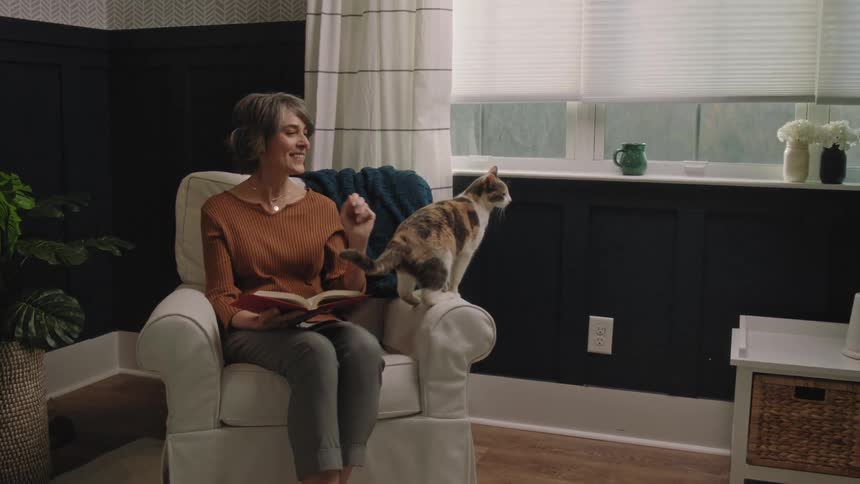 Woman sitting in a chair with a book in her lap watches her cat jump across her on the chair and reaches out to pet it